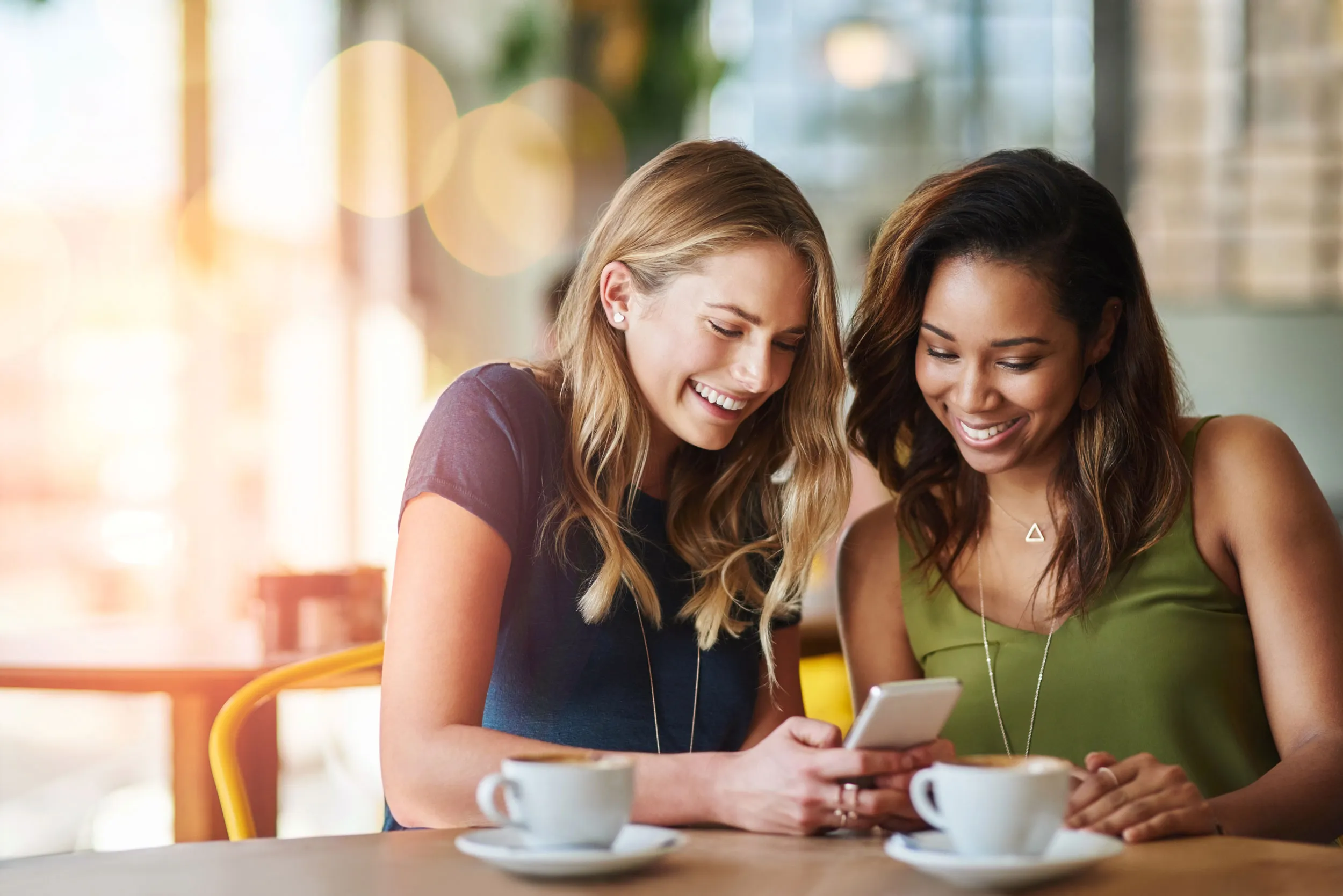 two woman are looking at a cell phone while sitting at a table with coffee